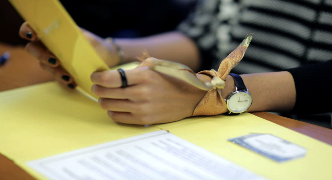 A person with a watch and a gold ribbon on their wrist writes on yellow paper at a desk, reflecting on understanding marginalized communities, with documents and a nametag nearby.