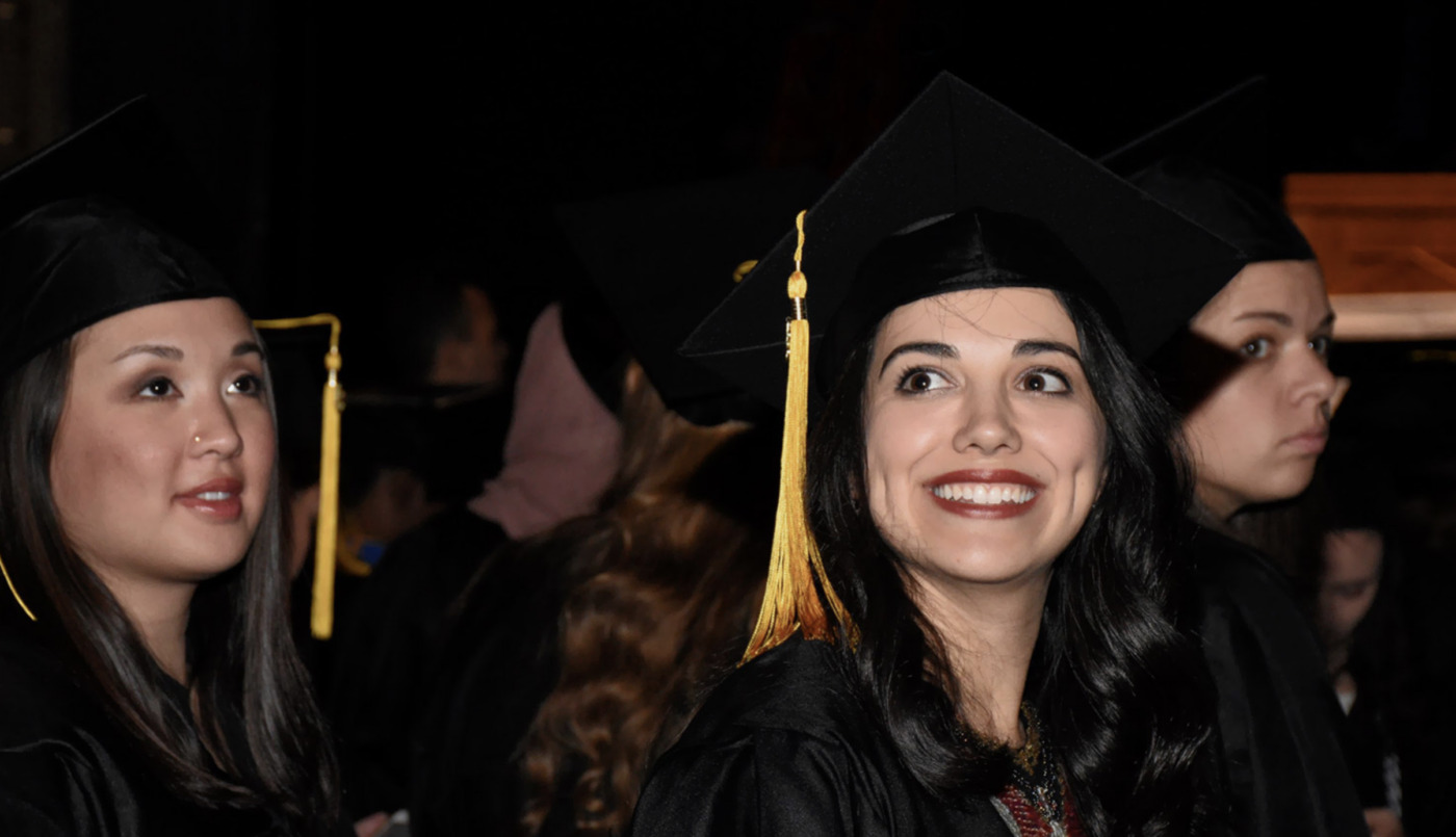 Three graduates in caps and gowns stand together indoors, facing forward; one woman in the center is smiling broadly, celebrating increased access to education.