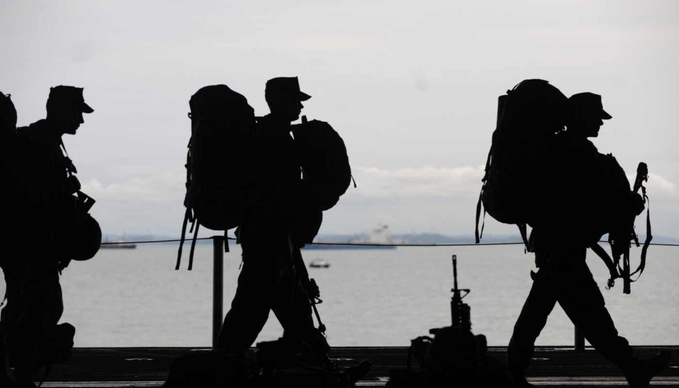 Silhouetted soldiers carrying backpacks and gear walk along a shoreline, water and boats in the background—a powerful reminder of the importance of veteran's mental health and ongoing mental health support for those who serve.