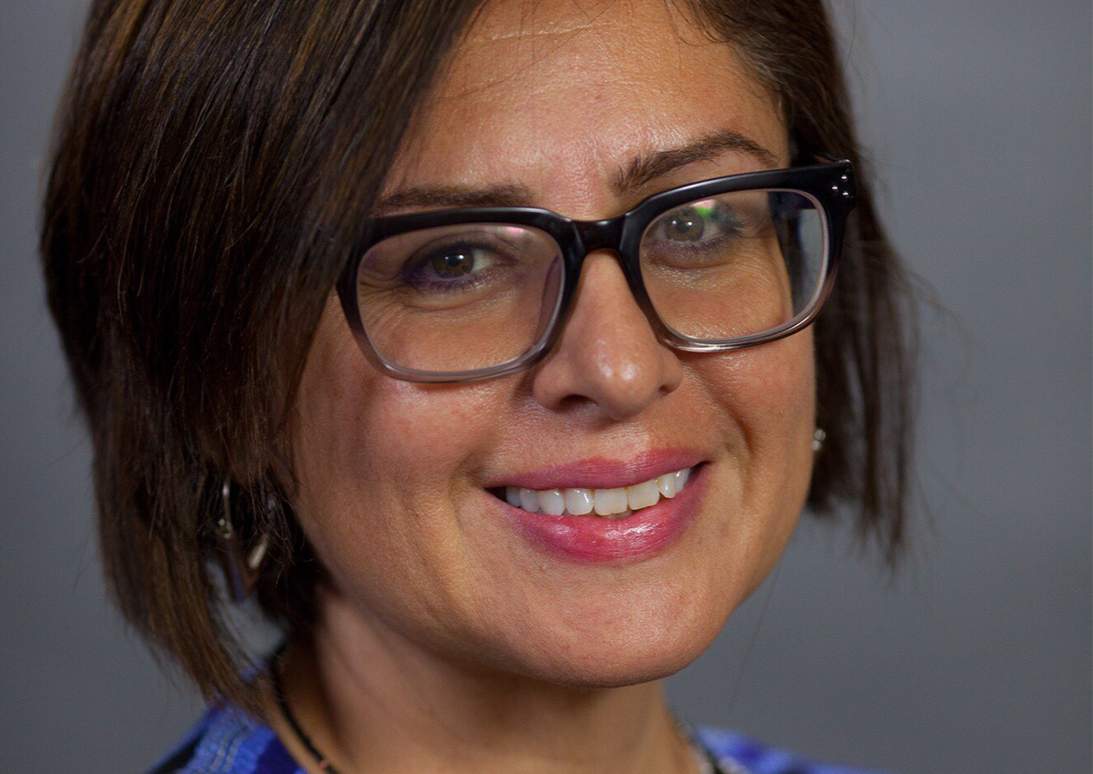 Shirley Aldana, a Change Agent with short brown hair and glasses, smiles at the camera in a blue top and light pink lipstick, set against a plain gray background.