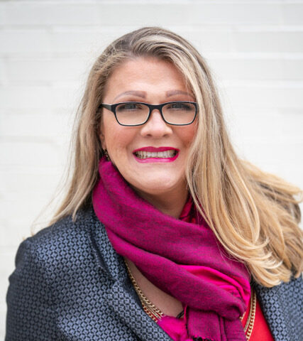 Dr. Stacee Reicherzer, with long blonde hair and glasses, smiles in a patterned blazer, bright pink scarf, and necklace, standing in front of a white brick wall—author of the handbook to heal otherness.