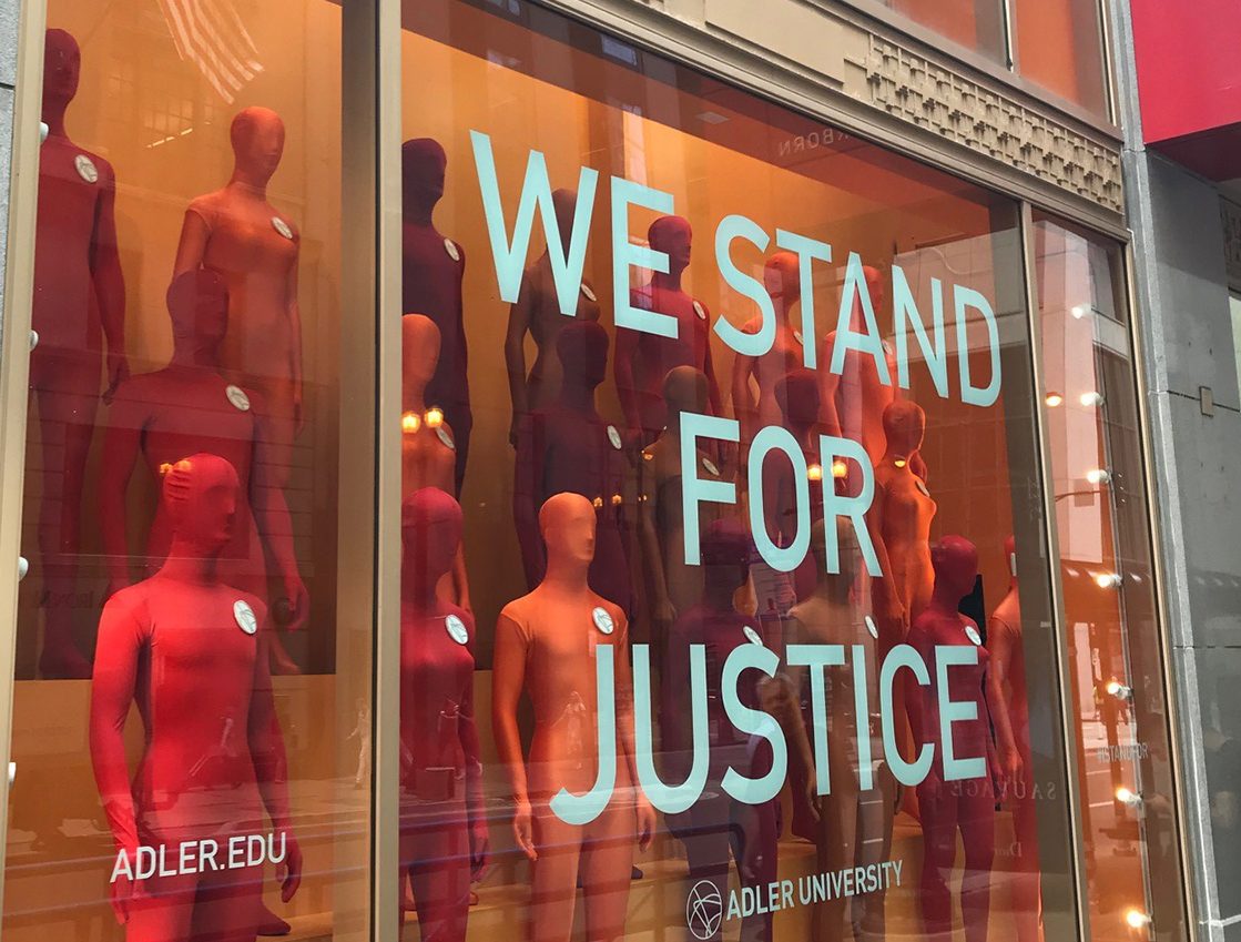 Storefront display with numerous red mannequins and large text on the window reading "WE STAND FOR JUSTICE." Adler University branding appears on the window.