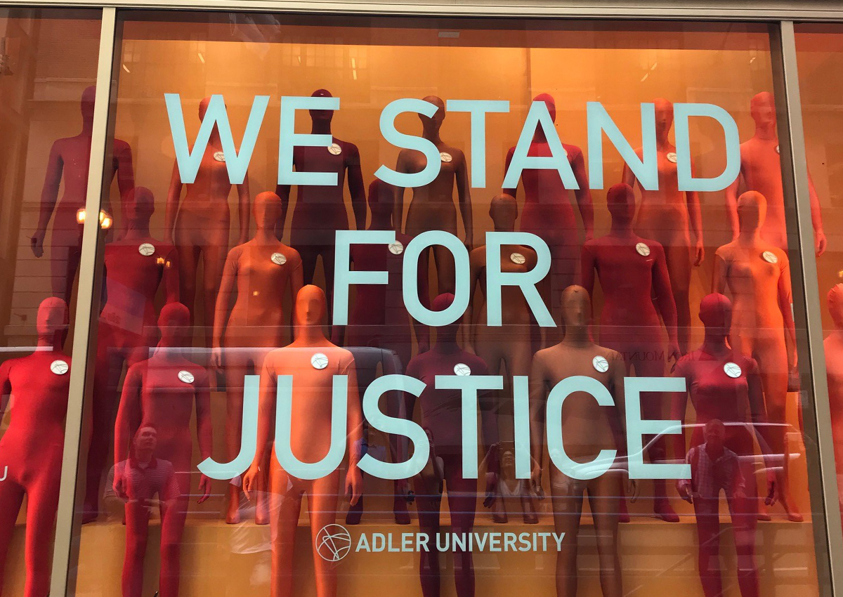 Window display featuring red and orange mannequins behind bold "We Stand For Justice" text and the Adler University logo, highlighting themes from the Social Justice Summit.