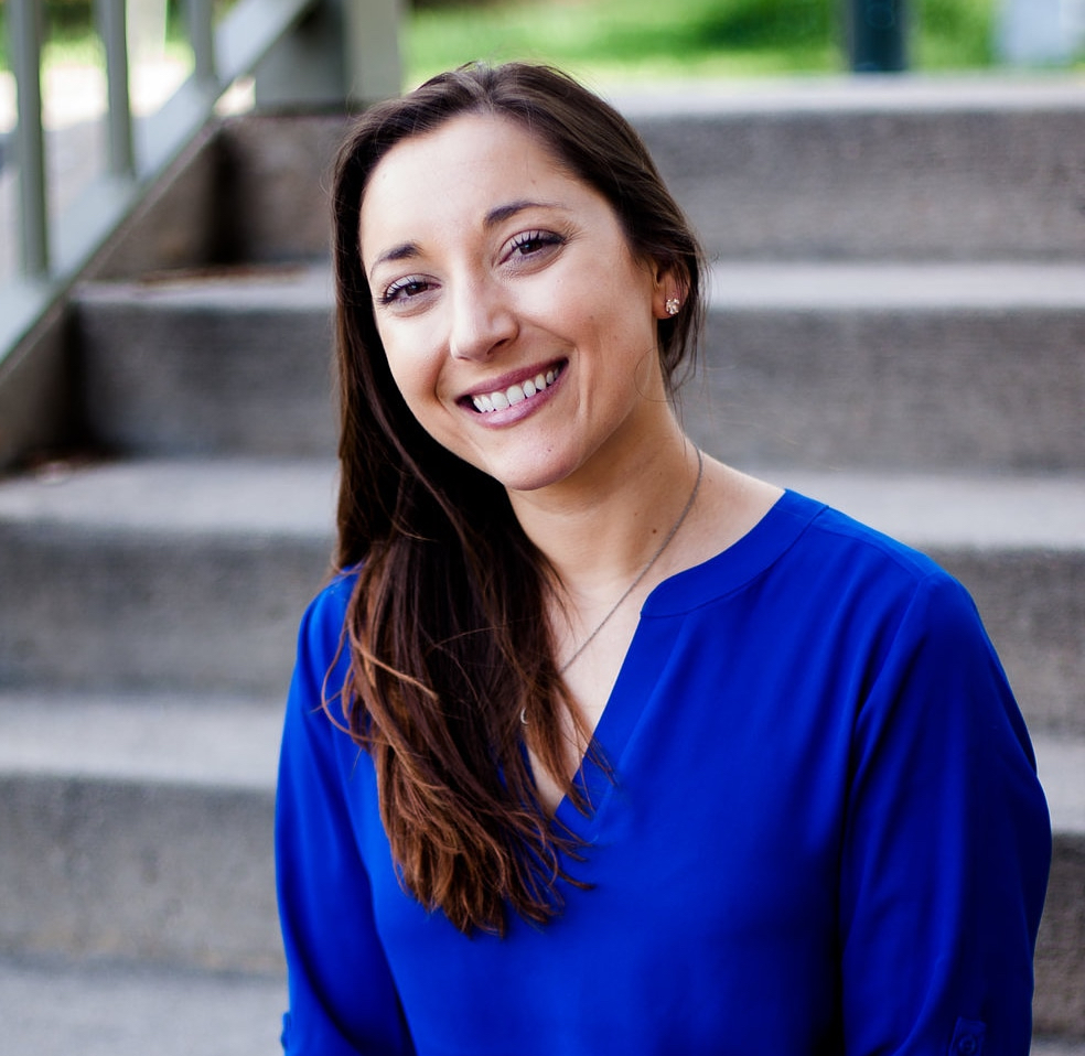 Woman with long brown hair in a blue blouse sits on outdoor steps, smiling at the camera—her work co-responding with law enforcement during mental health crises highlights her commitment to community well-being.
