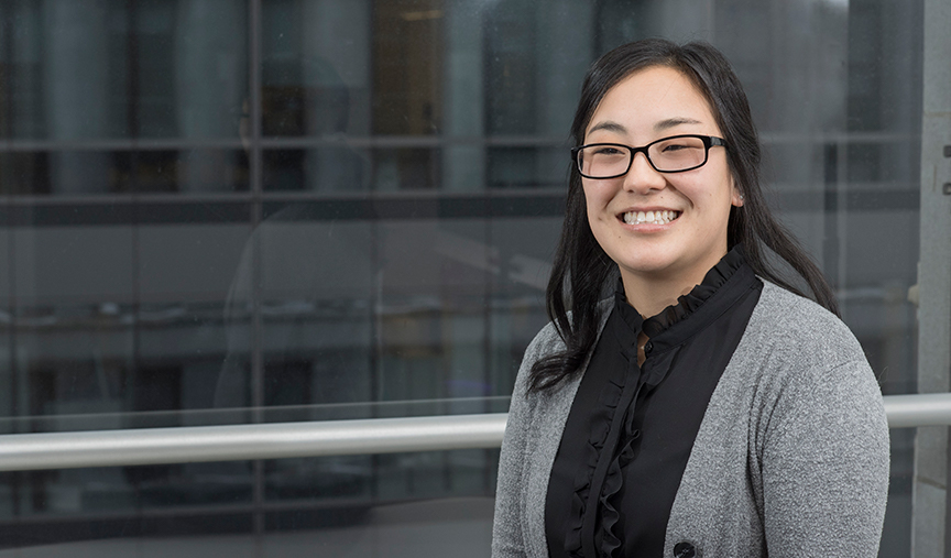 A woman wearing glasses, a black blouse, and a gray cardigan is smiling while standing in front of a window with an urban cityscape background.