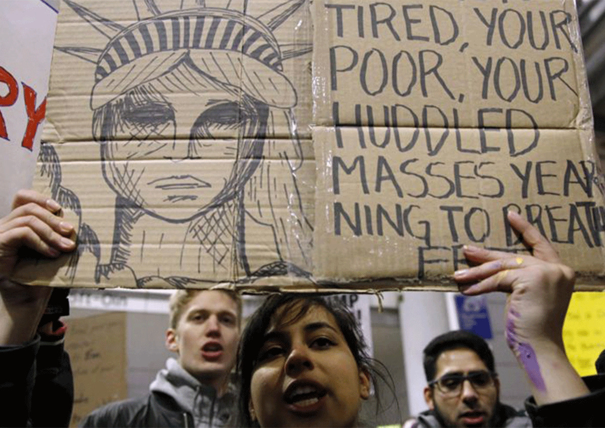 Protesters hold a cardboard sign with a drawing of the Statue of Liberty and handwritten text quoting "Give me your tired, your poor," calling for justice and compassion in the administration of immigration policies.