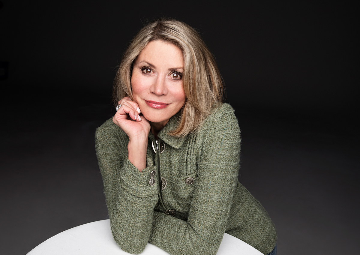 A woman with blonde hair wearing a green textured jacket rests her chin on her hand and looks at the camera, reflecting on community mental health and well-being against a dark background.