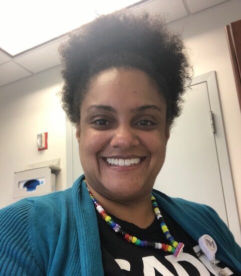 A woman with curly hair smiles at the camera, wearing a colorful beaded necklace, blue cardigan, and a black shirt indoors. She is the Director of Training for the ACHS Division of Community Health in Chicago.