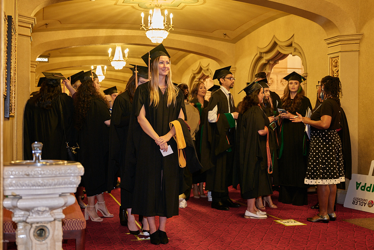 A group of graduates in caps and gowns stand and converse in a hallway with arched doorways and chandeliers.