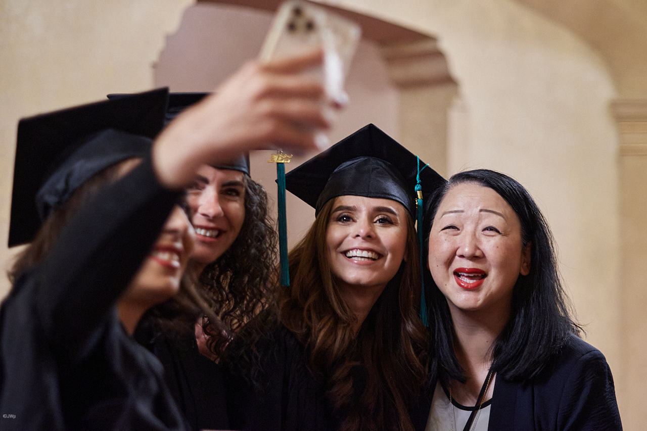 Four women in graduation caps and gowns smile as they take a selfie together indoors.