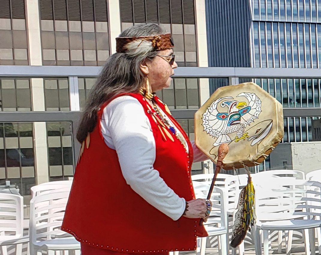A person wearing a red vest and headband stands outdoors holding a painted ceremonial drum, symbolizing reconciliation, with city buildings and stacked white chairs in the background.