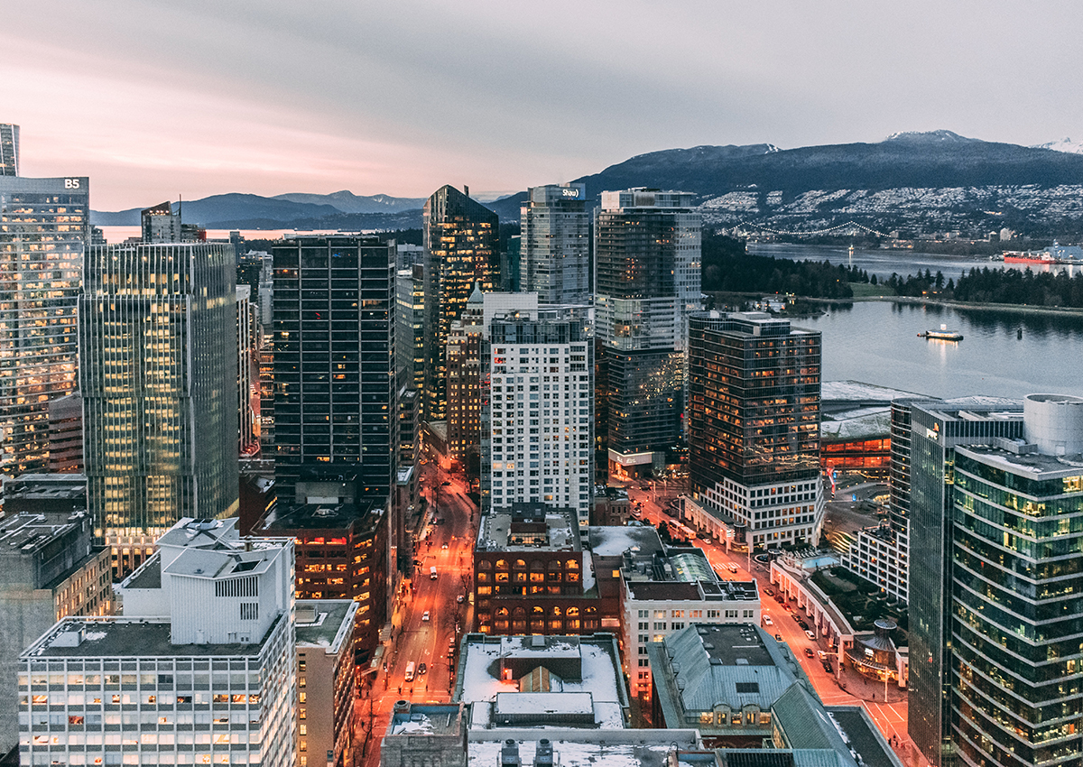 Aerial view of a modern city with tall buildings, illuminated streets, and a harbor with mountains in the background during dusk, home to initiatives like Vancouver Foundation’s Emergency Community Support Funds.