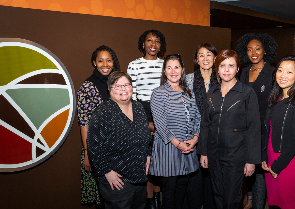 A group of eight women, possibly students, pose together indoors near a circular, multicolored abstract wall logo—celebrating International Women’s Day.