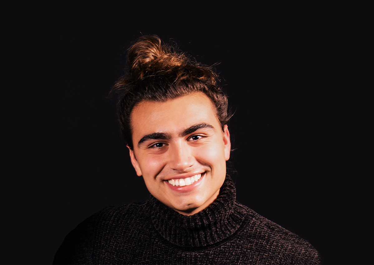 A person with long dark hair in a top bun, wearing a black turtleneck sweater, smiles against a plain black background—capturing the quiet strength often seen in LGBTQ+ Art Therapists and those championing Invisible Histories through Art Therapy.