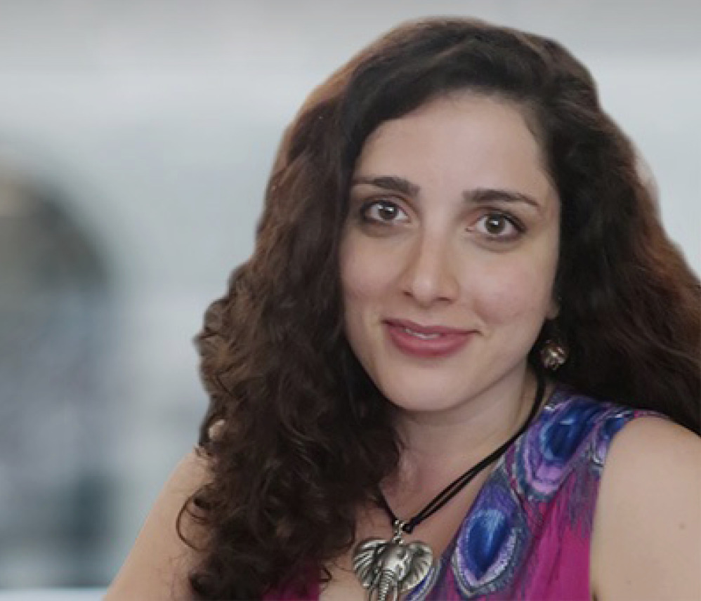 A woman with long curly hair, wearing a colorful top and a large heart-shaped pendant, is sitting indoors and looking at the camera, embodying mental wellness and positivity.