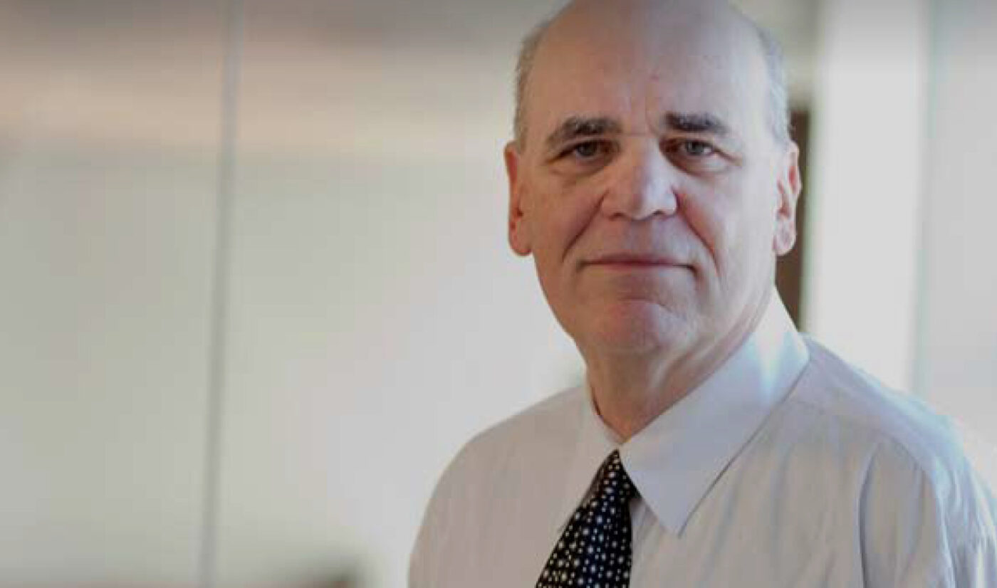An older man with a receding hairline and white shirt with a black patterned tie stands indoors, facing the camera—his expression reflecting the challenges of poverty and the importance of education in El Salvador Stories.
