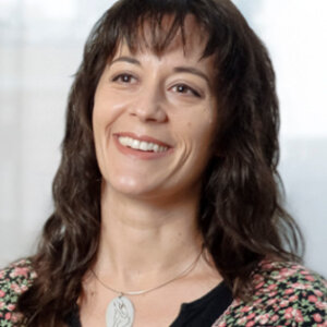 Dr. Maglio is standing in front of a large window with a building blurred in the background. She is wearing a blouse.