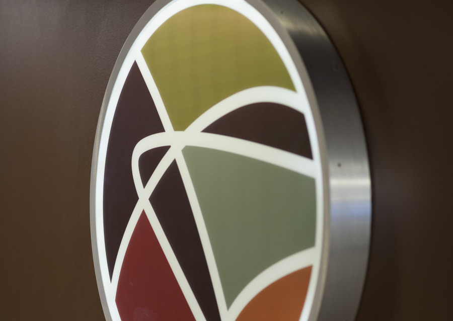 A round wall sign with geometric shapes in green, red, yellow, and gray, outlined in white, mounted on a brown background.