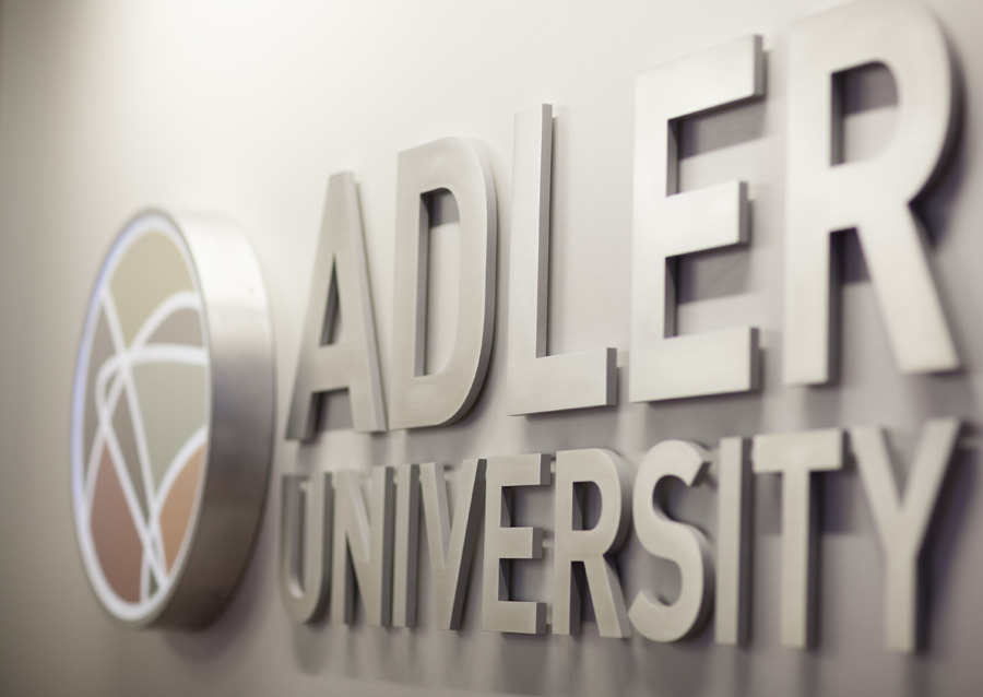 A silver sign on a wall displays the Adler University logo and name in large, raised letters, welcoming students for the Spring Term of virtual instruction.