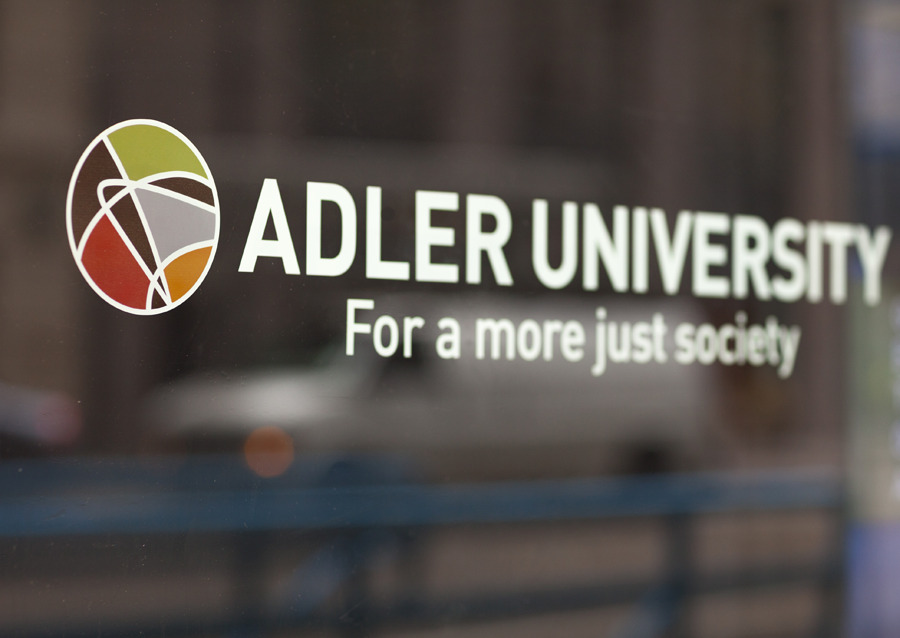 Adler University logo and slogan “For a more just society” displayed on a glass window, with a blurred street scene reflected in the background.