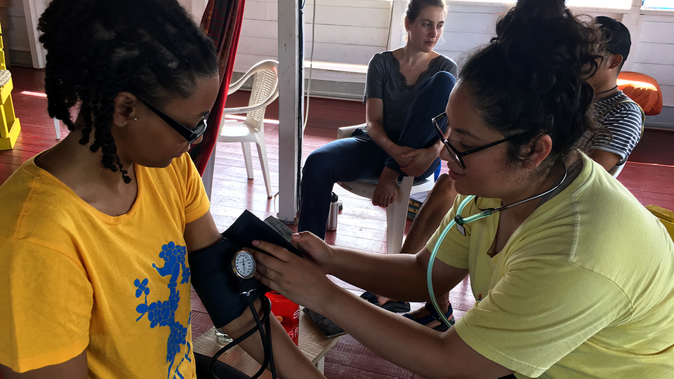 A healthcare worker checks another person’s blood pressure while two others observe in a room with red flooring and white walls, highlighting the importance of increasing access to mental health care.