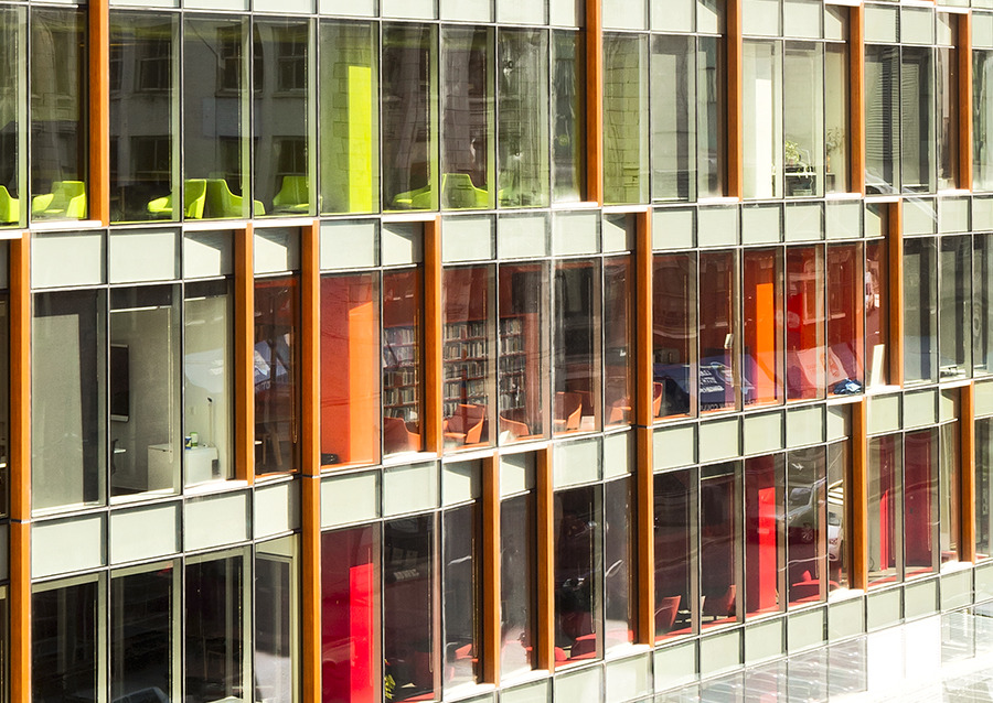 A modern building facade with large glass windows, some tinted orange or red; green and orange chairs are visible inside through the windows.