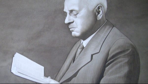 A grayscale portrait of an older man in a suit and glasses, holding and reading an open book.