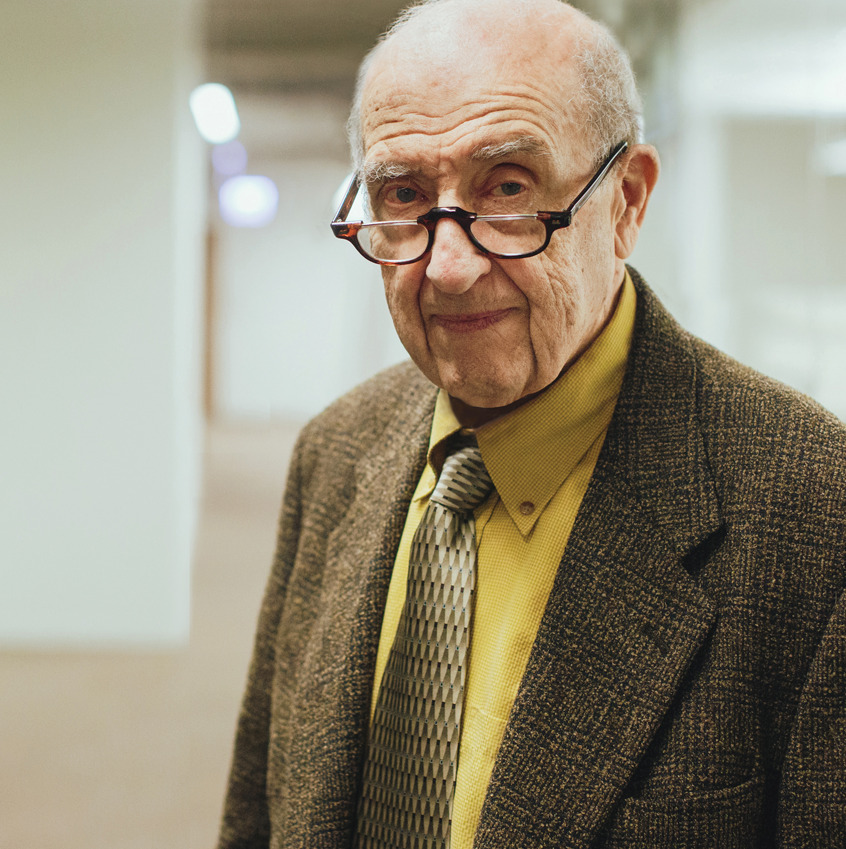 Dr. Bernard Shulman, an elderly man in glasses, a brown tweed jacket, yellow shirt, and patterned tie, stands indoors looking at the camera.