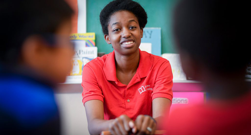 A young woman in a red polo shirt, embodying a true Change Agent, sits at a desk in a classroom, facing two students who are out of focus in the foreground.