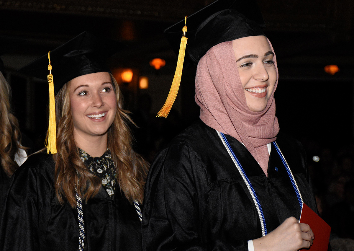 Two 2019 graduates wearing caps and gowns stand side by side, smiling during a Vancouver graduation ceremony. One wears a hijab and holds a diploma, reflecting the city’s diversity and spirit of social justice.