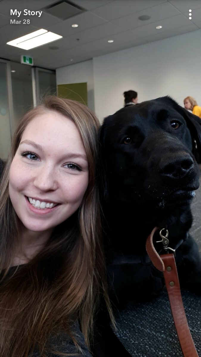 A woman smiles beside a black dog wearing a leash indoors, with other people visible in the background, highlighting the benefits of pet therapy in bringing joy and connection to group settings.