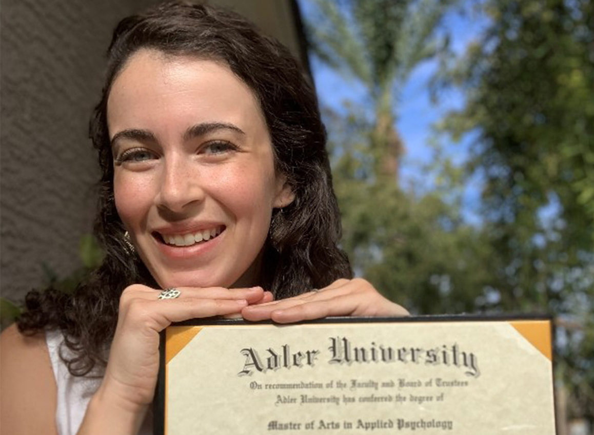 A smiling online graduate holds an Adler University Master of Arts in Applied Psychology diploma outdoors, with trees symbolizing the seeds of change they've planted through their education.