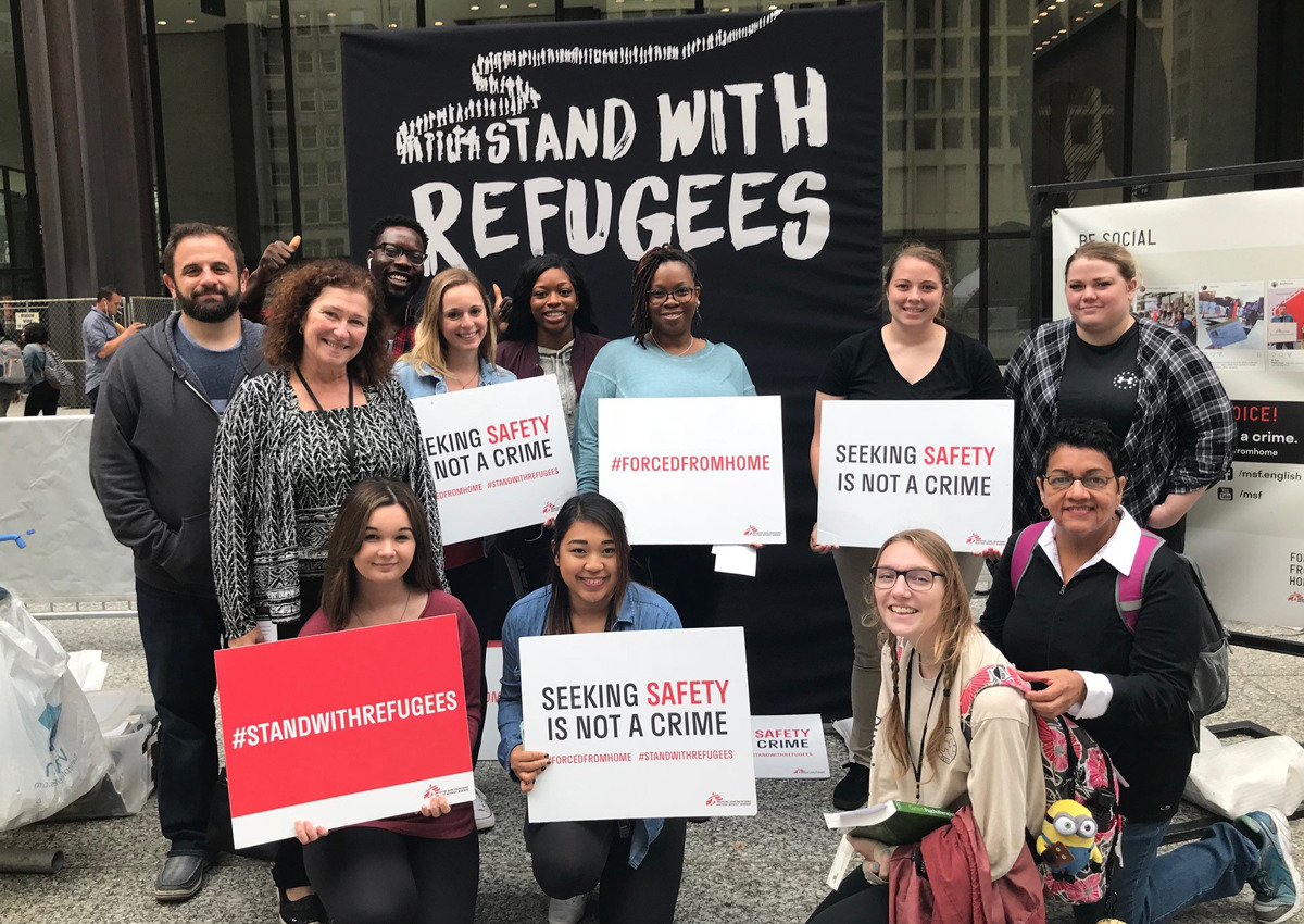 A group of people pose in front of a "Stand With Refugees" banner, holding signs that say "Seeking safety is not a crime" and "#StandWithRefugees," raising awareness about the global refugee crisis and the effects of displacement.