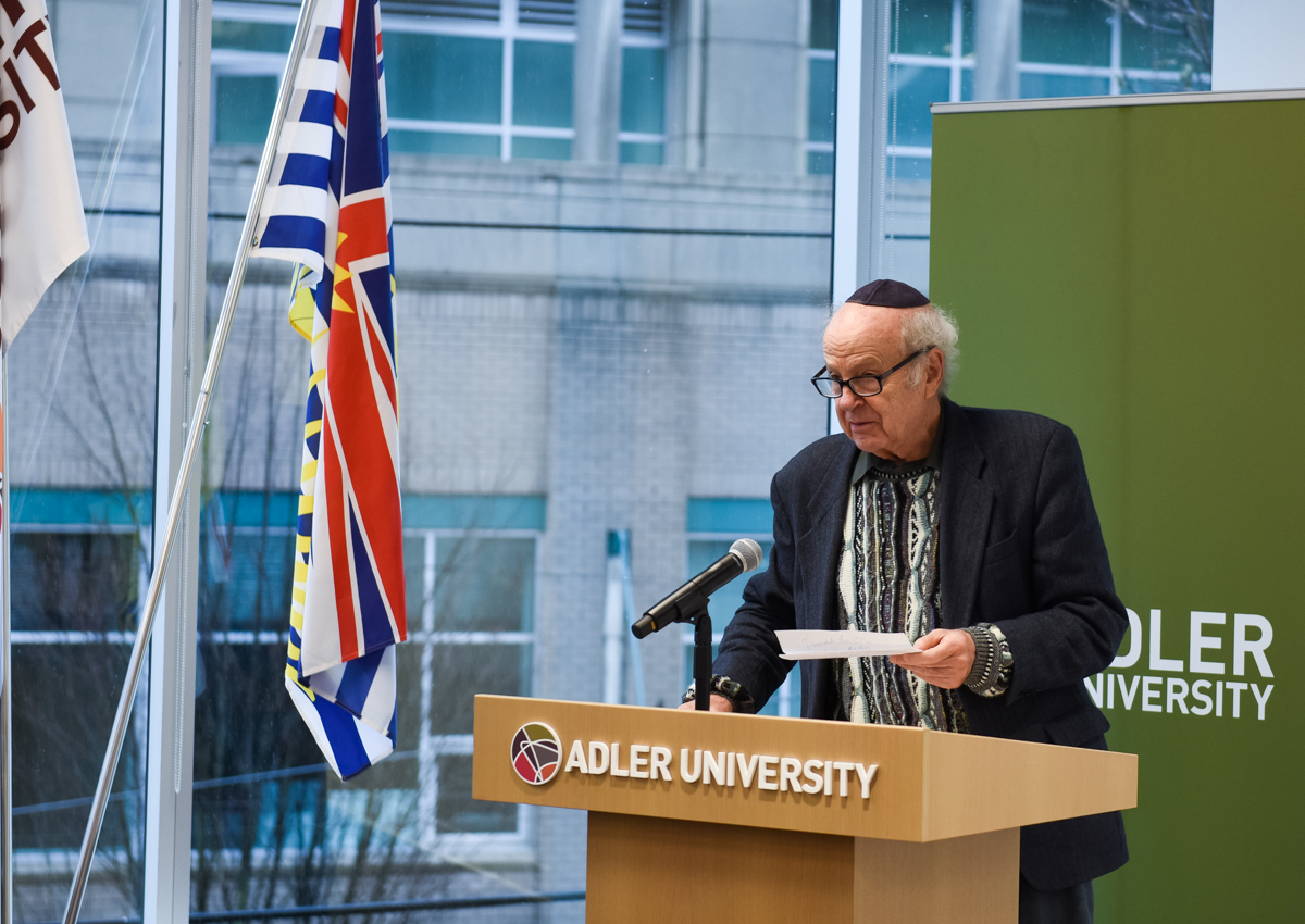 An older man wearing a kippah speaks at a podium labeled "Adler University," with flags and a green Adler University banner in the background, during a Vancouver event celebrating an Endowed Scholarship in Counselling Psychology.