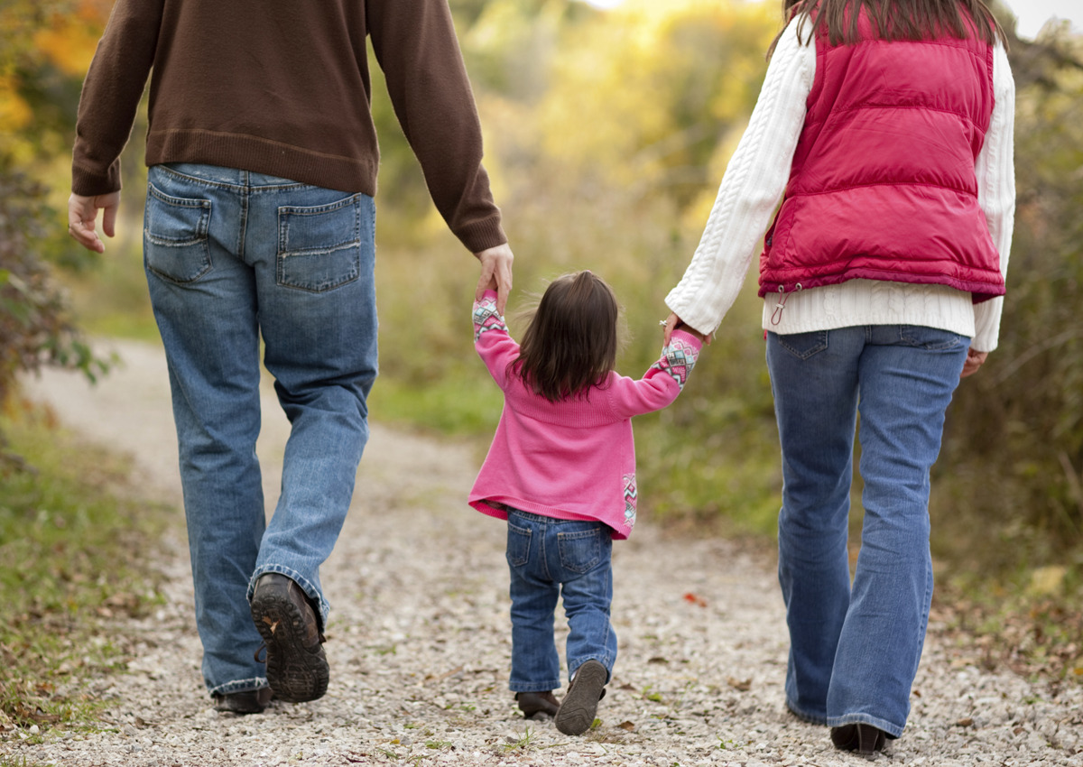 Two adults and a child walk hand-in-hand on a gravel path outdoors, photographed from behind. This warm moment highlights the family bonds often explored in Couple and Family Therapy Ph.D. programs with national accreditation.