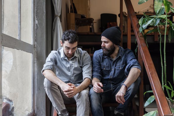 Two men sit on stairs indoors, one looking down with hands clasped, the other looking at him, appearing to have a serious conversation.