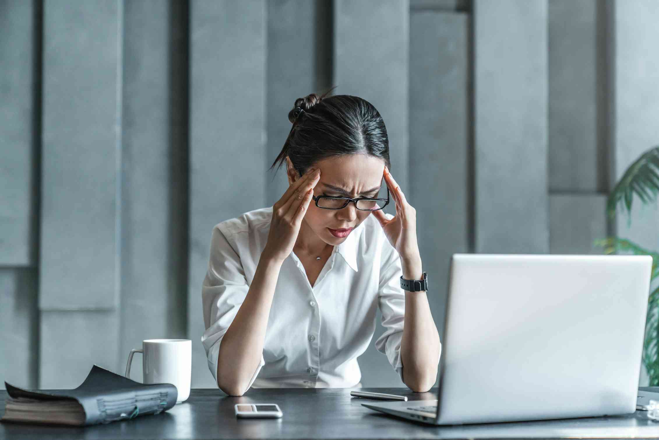 A woman in a white shirt sits at a desk with a laptop, holding her temples and appearing stressed or frustrated.
