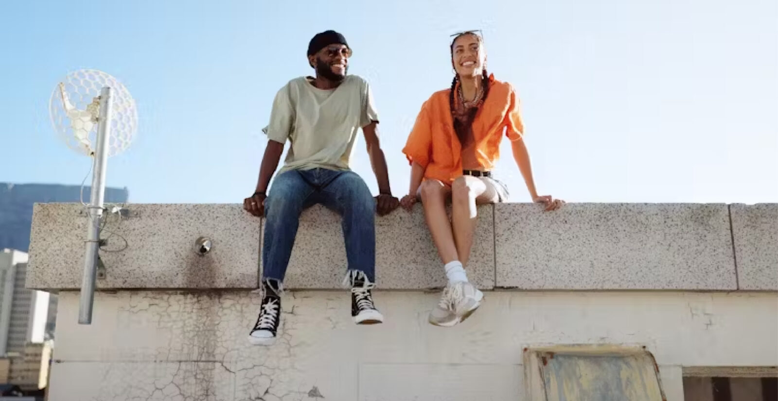 Two people sit on a rooftop ledge in casual clothes, smiling at each other under a clear sky with city buildings behind them, capturing a moment of authentic relationships.