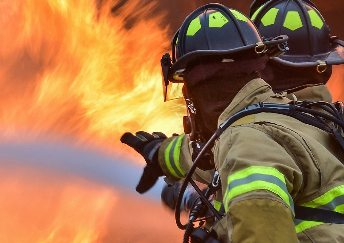 Two first responders in protective gear use a hose to fight a large blaze, with bright flames visible in the background—highlighting both their bravery and the critical need for mental health care support.