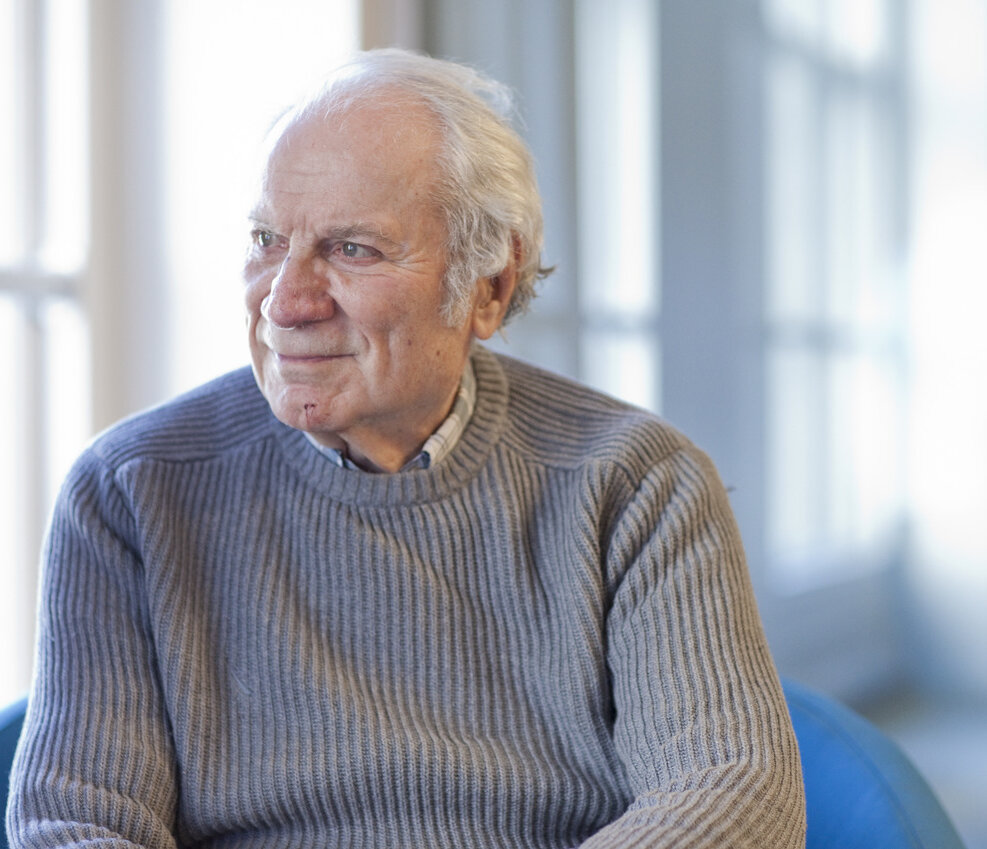 In Memoriam: Dr. Harold Mosak, an elderly man with white hair, sits on a blue chair in a gray ribbed sweater, looking to his left with a slight smile as natural light softly illuminates the background.