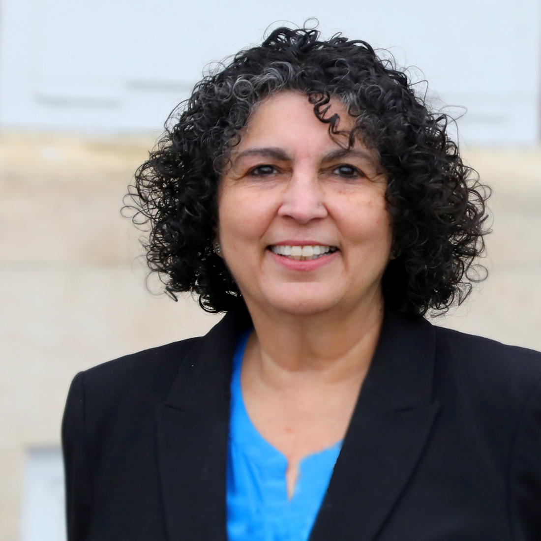 Rita M. Dibble, vice president of institutional advancement at Adler University, with curly dark hair and some gray, wearing a black blazer over a blue blouse, stands outdoors and smiles at the camera.