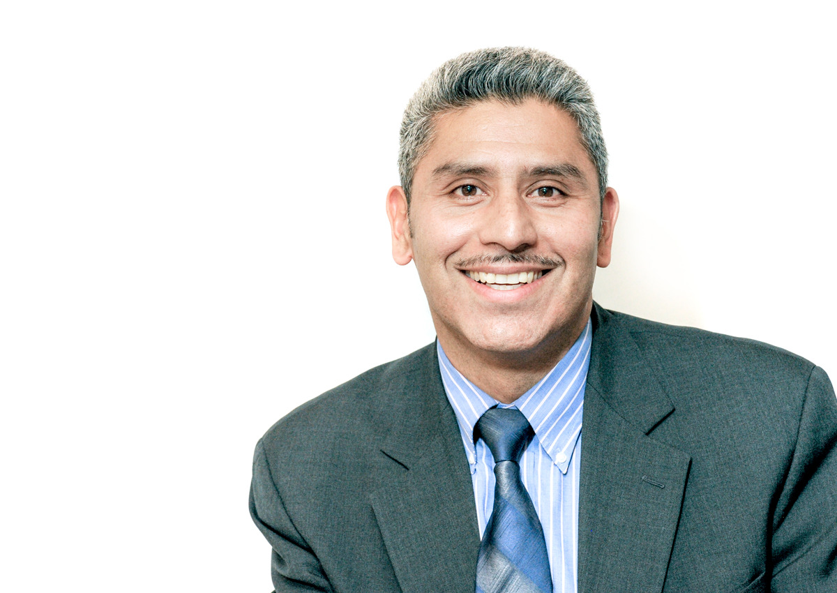 A man in a suit and tie with short gray hair and a mustache smiles at the camera against a plain white background, embodying professionalism in the field of education and its social determinants.