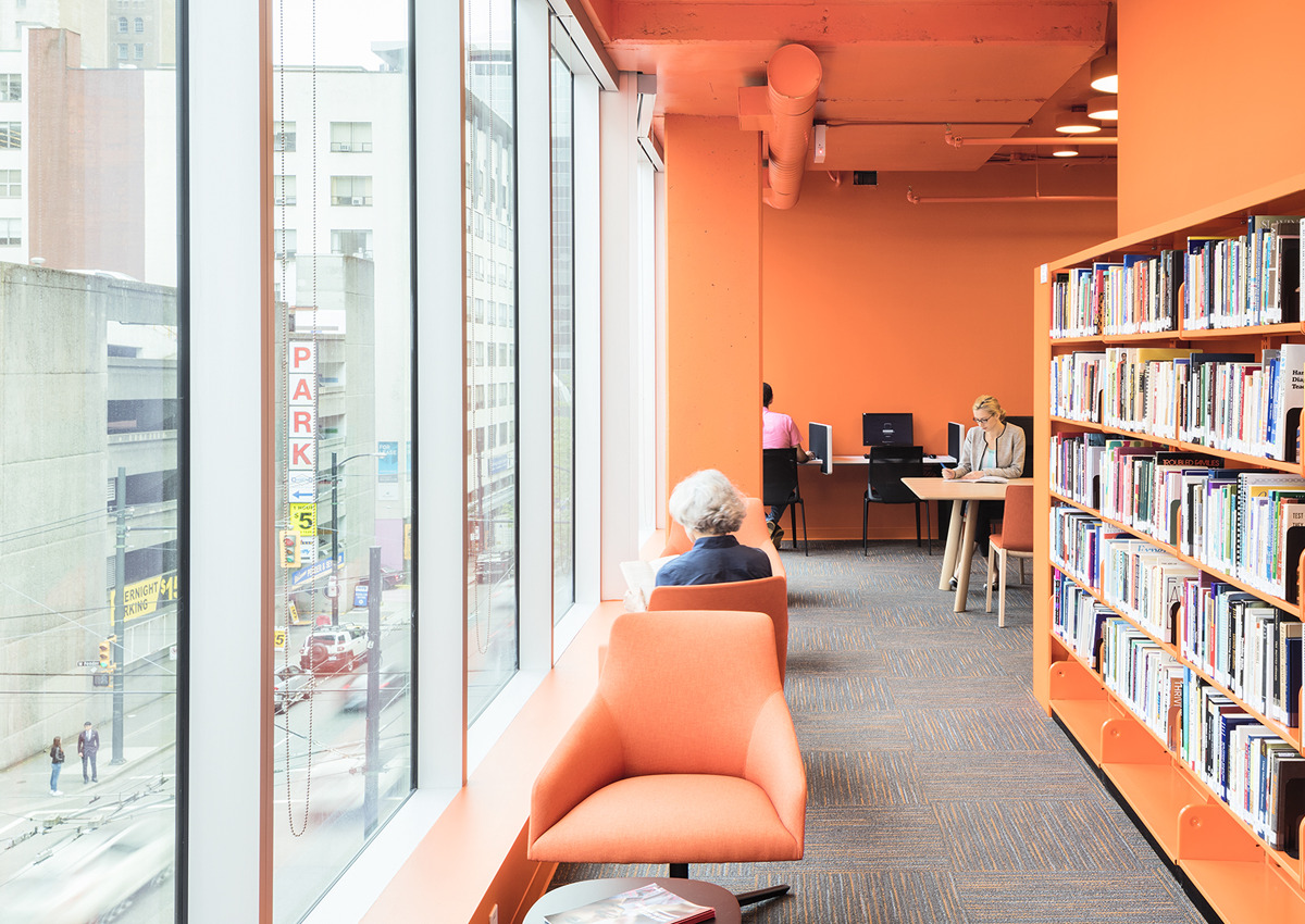 A modern library interior with orange walls and chairs, large windows overlooking a city street, bookshelves, and people reading or working on computers.
