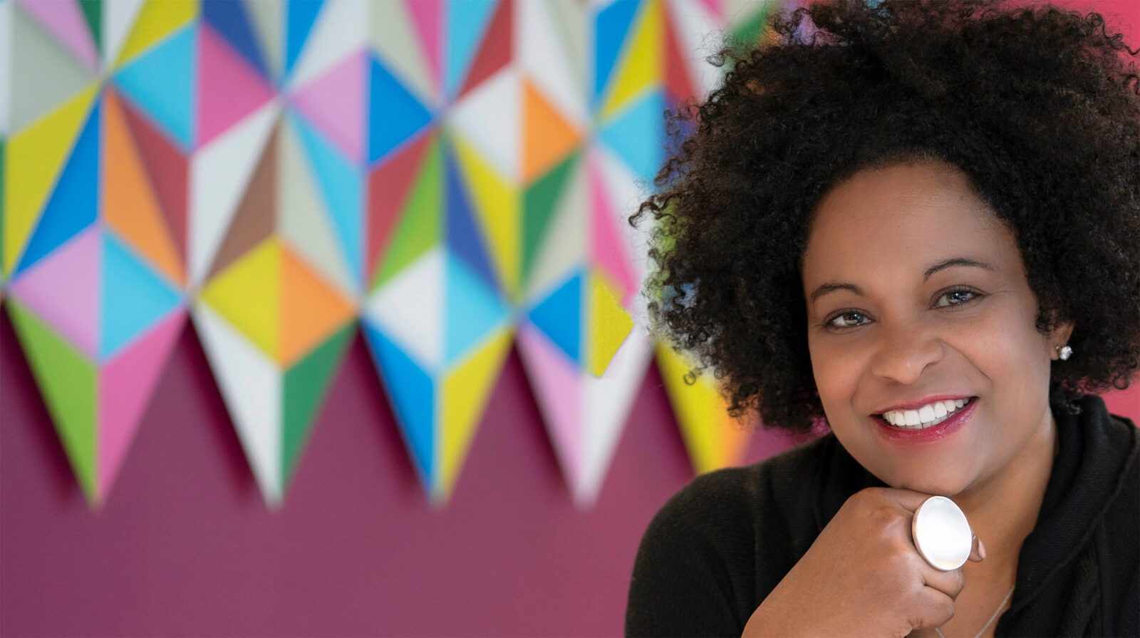 A woman with curly hair smiles at the camera, resting her chin on her hand. Adler President Lisa Coleman, a Top Women Leader, is pictured with a colorful geometric pattern displayed on the wall behind her.