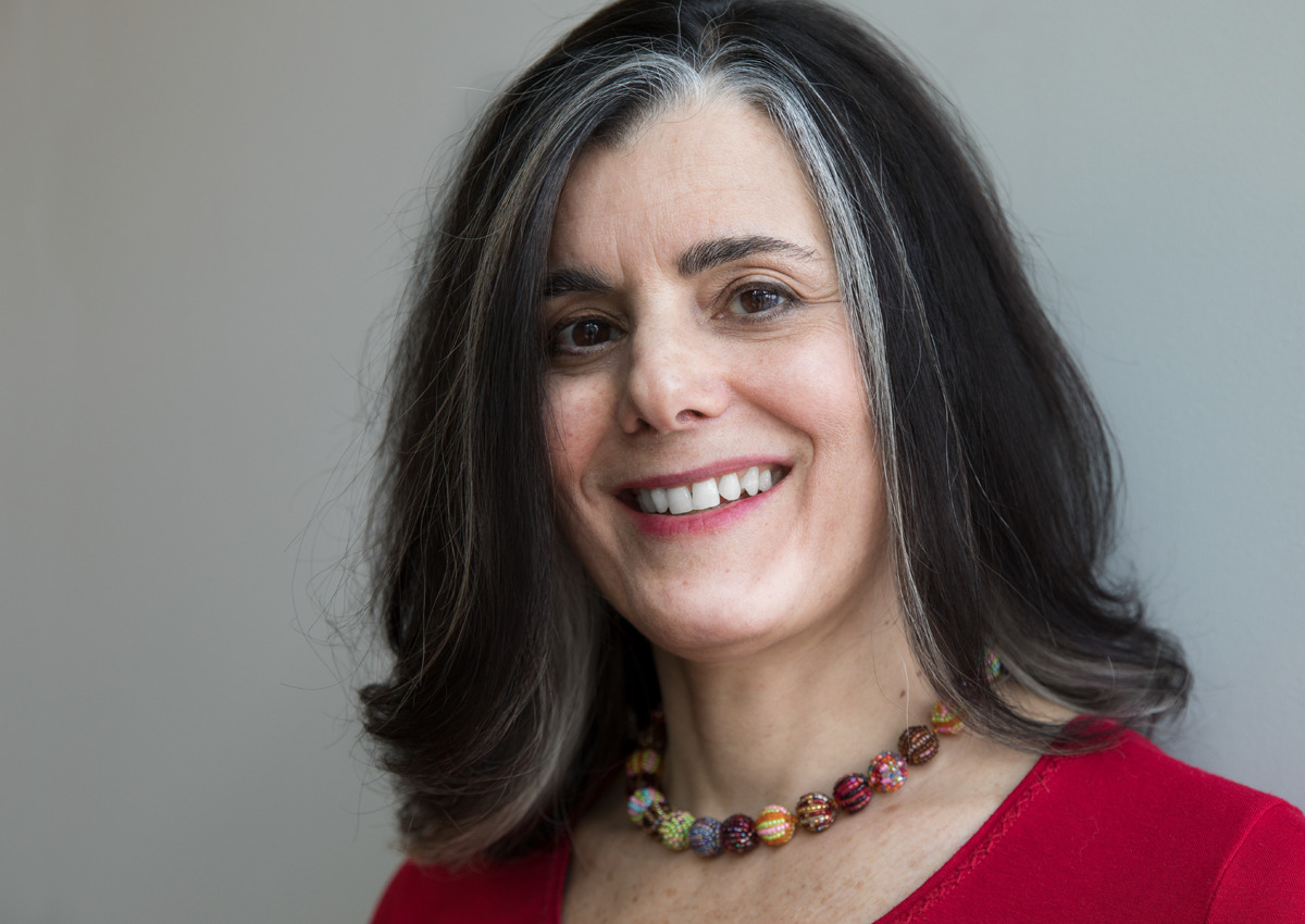A woman with shoulder-length dark hair streaked with gray, a red top, and a colorful beaded necklace smiles at the camera; she is a Fellow of the American Council on Education at Adler University, posing against a plain background.