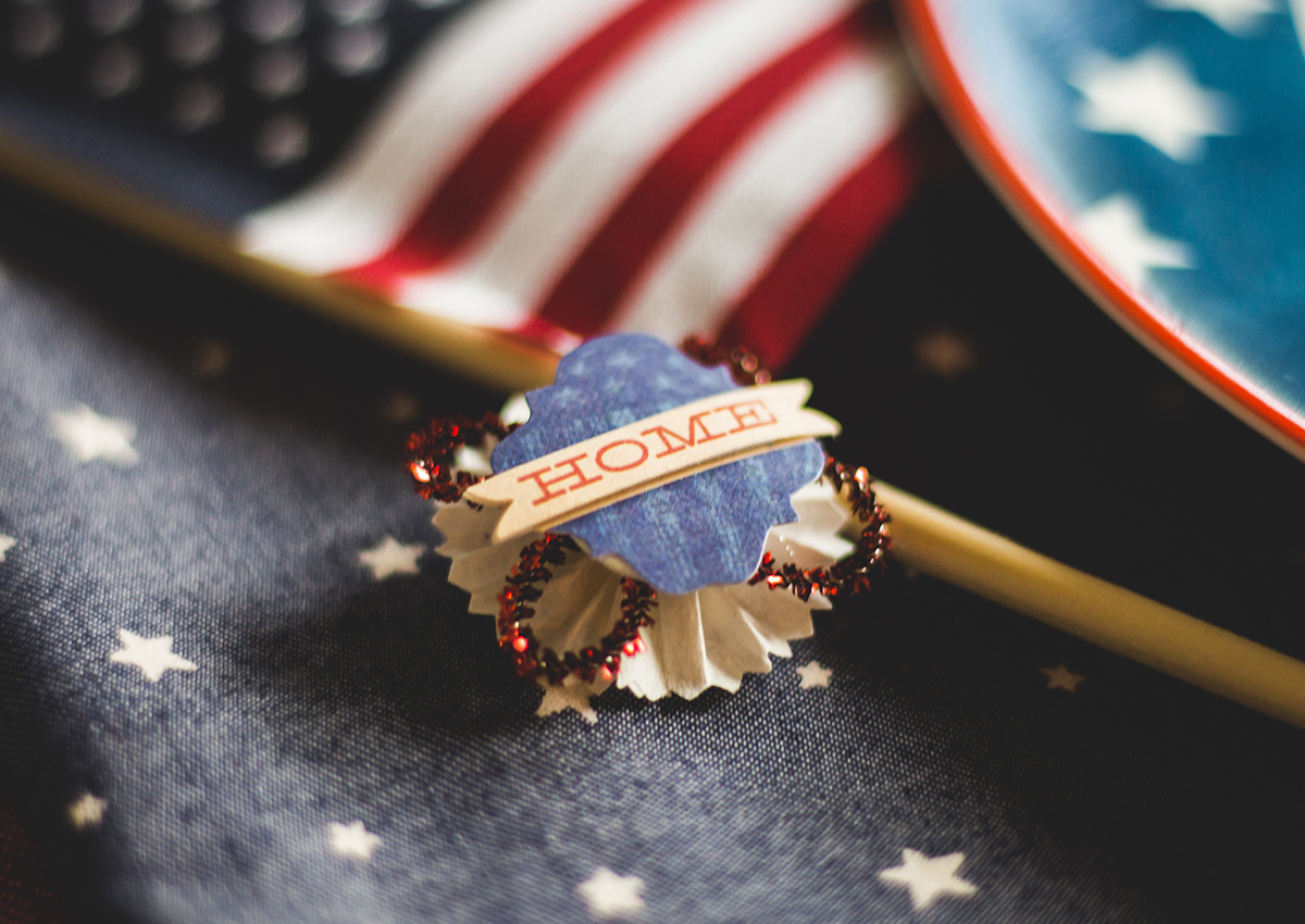 A decorative pin with the word "HOME" sits on star-patterned fabric, honoring Veterans, with part of an American flag and a blue plate with white stars in the background.
