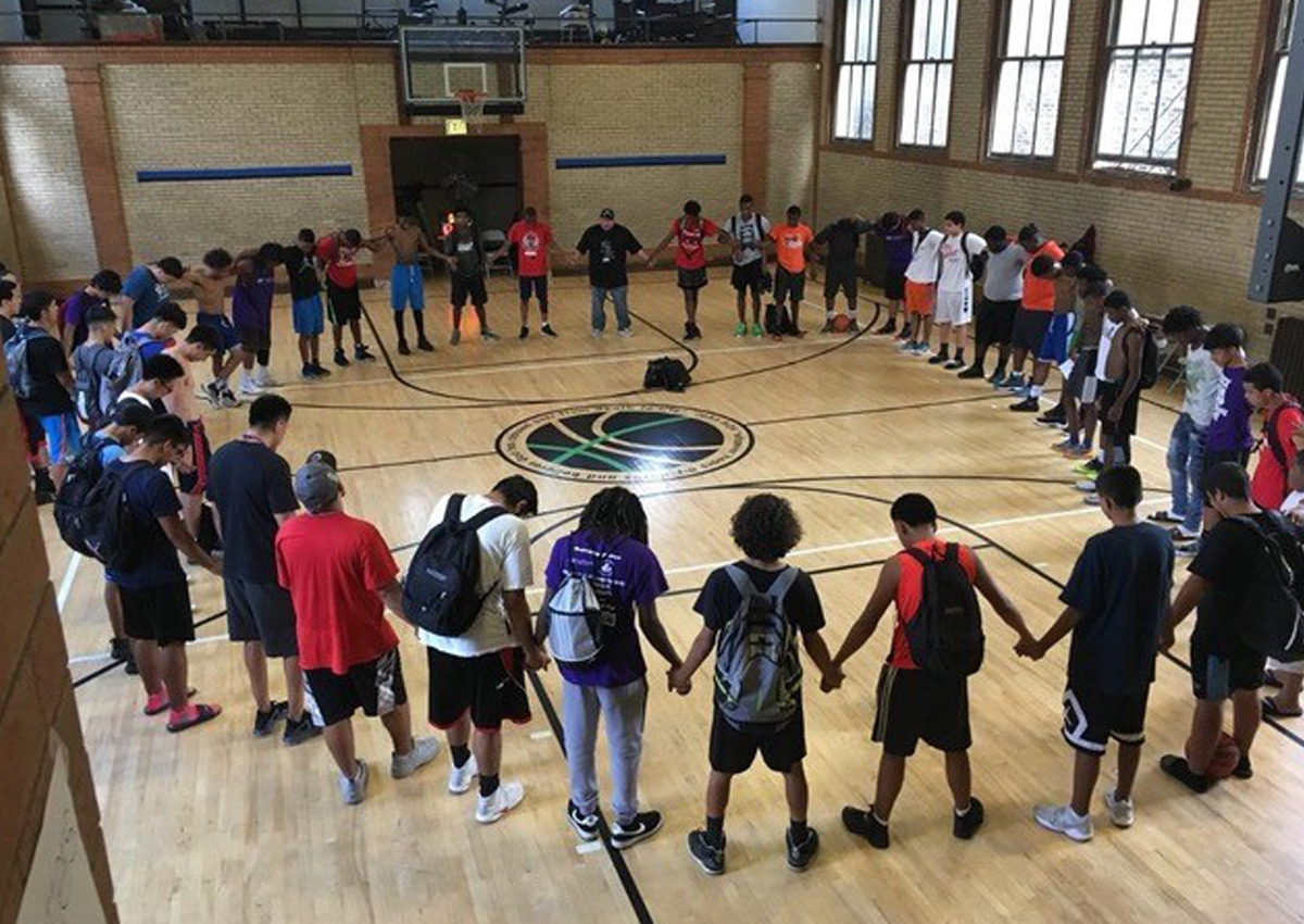 A group of people stand in a circle holding hands at the center of an indoor basketball court, united by community organizations and a shared commitment to restorative justice.