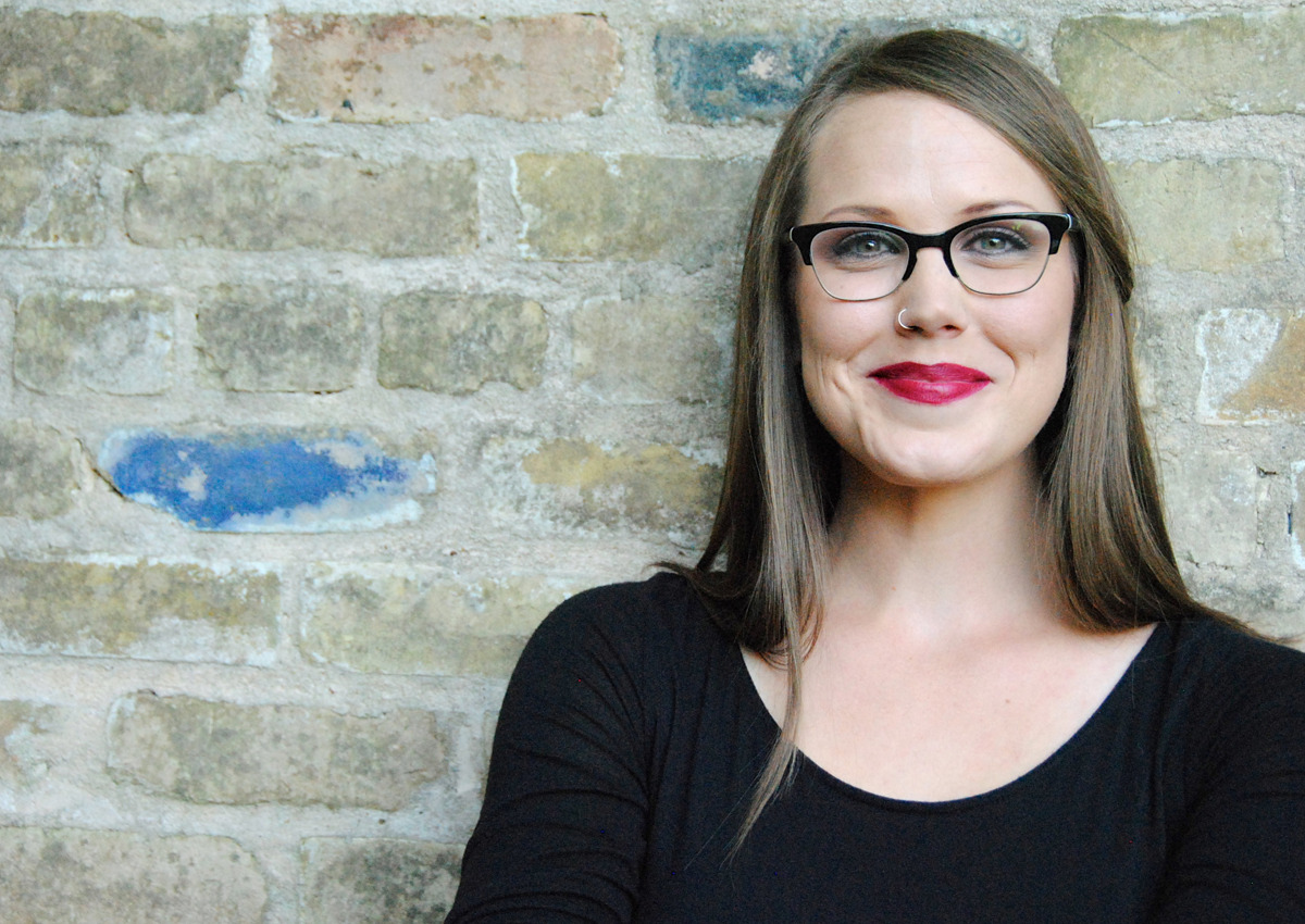 A woman with long brown hair and glasses, wearing a black top, stands in front of a brick wall and smiles at the camera, inspiring children to dream and love courageously.