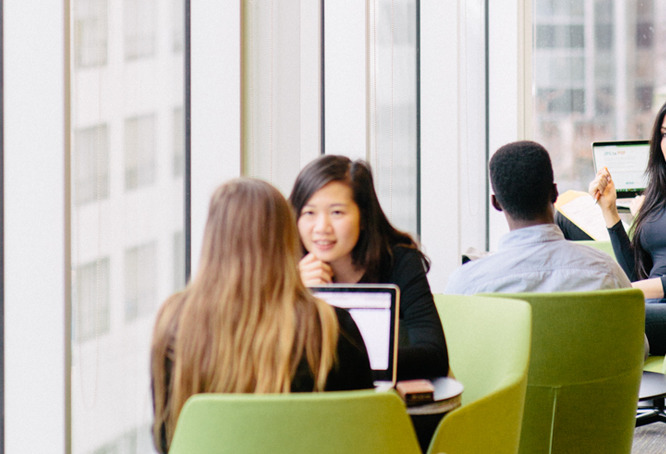 Two people talk across a table with laptops in front of them, while others sit nearby in green chairs by large windows in a modern office or study area.