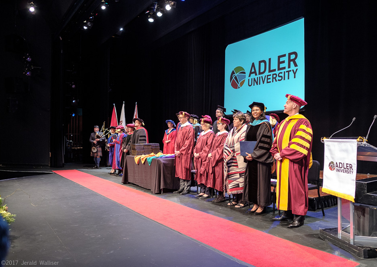 A group of faculty, officials, and 2018 graduates in academic regalia stand on stage at an Adler University graduation ceremony.